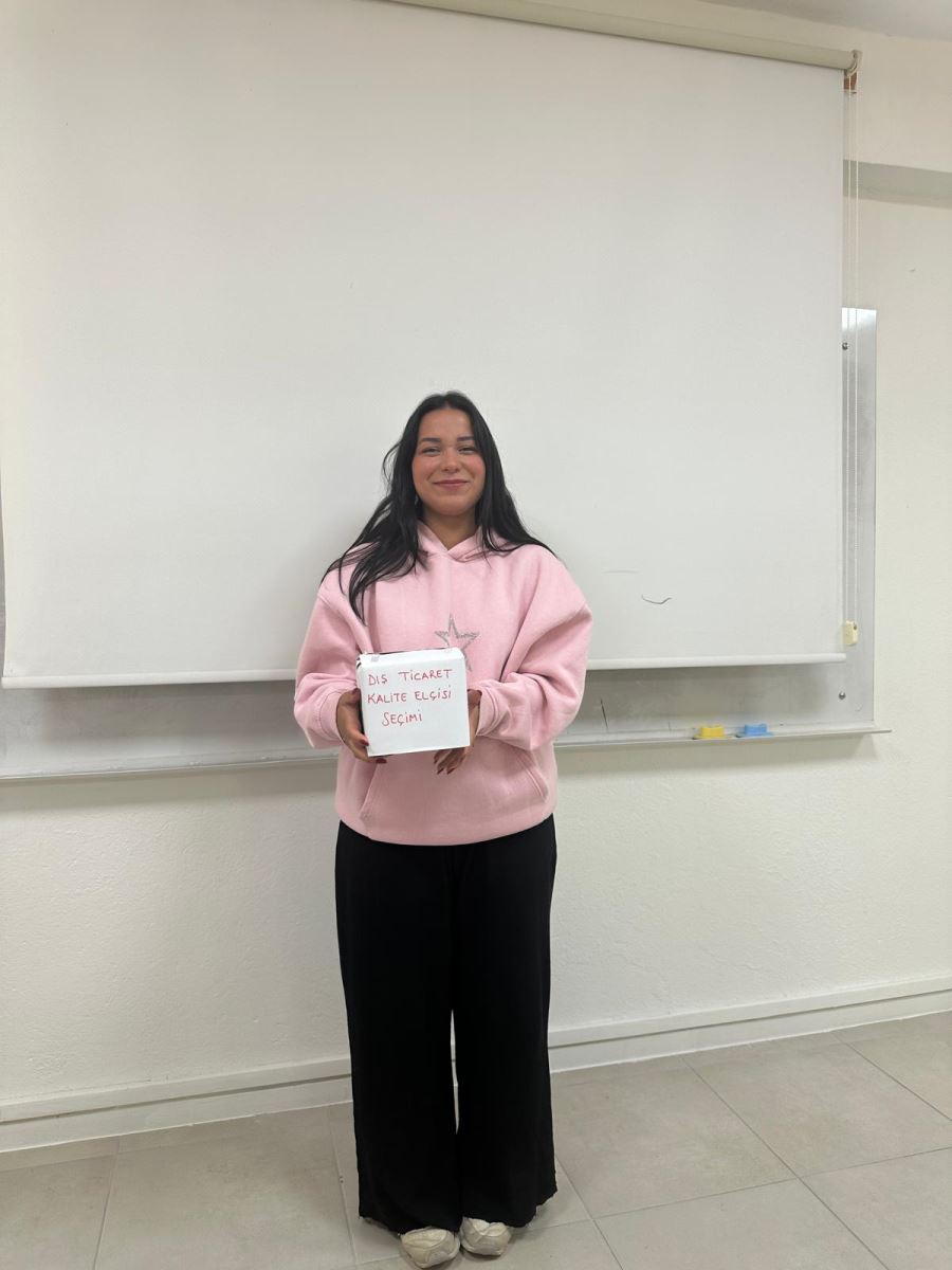 A single student stands alone in front of a white wall.  * She is wearing a light pink hoodie, loose black pants, and white sneakers.  * She is holding a white box with red writing that says “Foreign Trade Quality Ambassador Election.”  * She smiles gently at the camera, giving the photo a warm and cheerful feeling.
